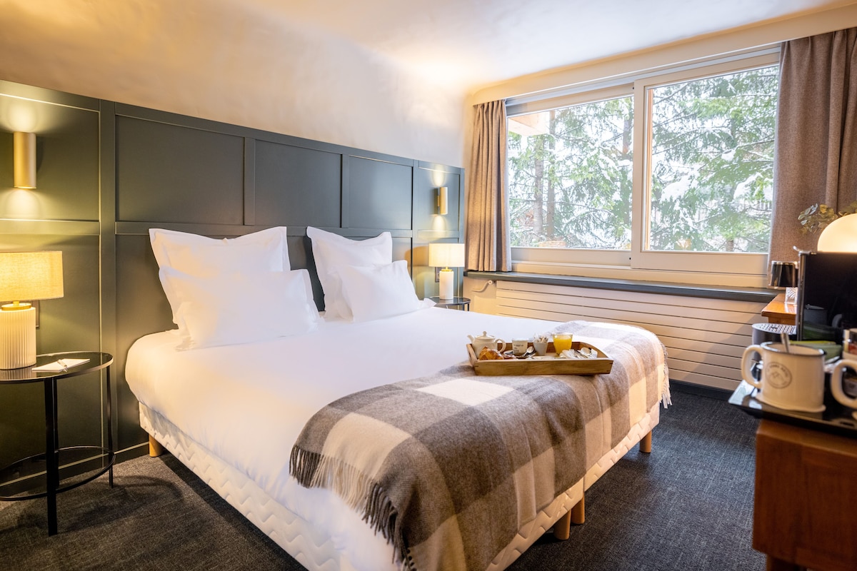 A hotel room with a large bed, a bedside table with a lamp, a tray with breakfast items, and a window with a view of trees.