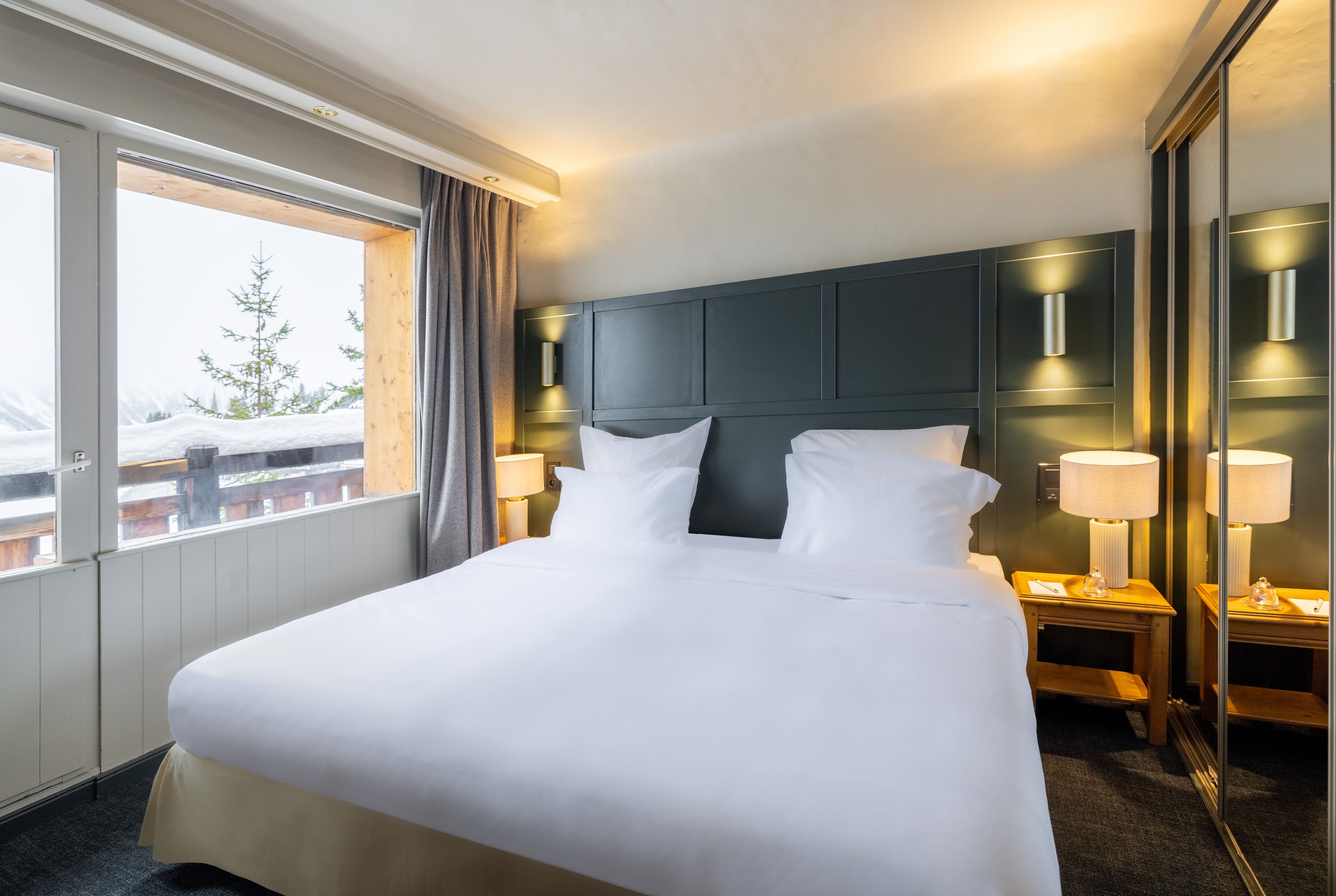 Une chambre d’hôtel avec un grand lit, deux lampes de chevet, une table de chevet, un miroir et une fenêtre donnant sur des arbres couverts de neige.