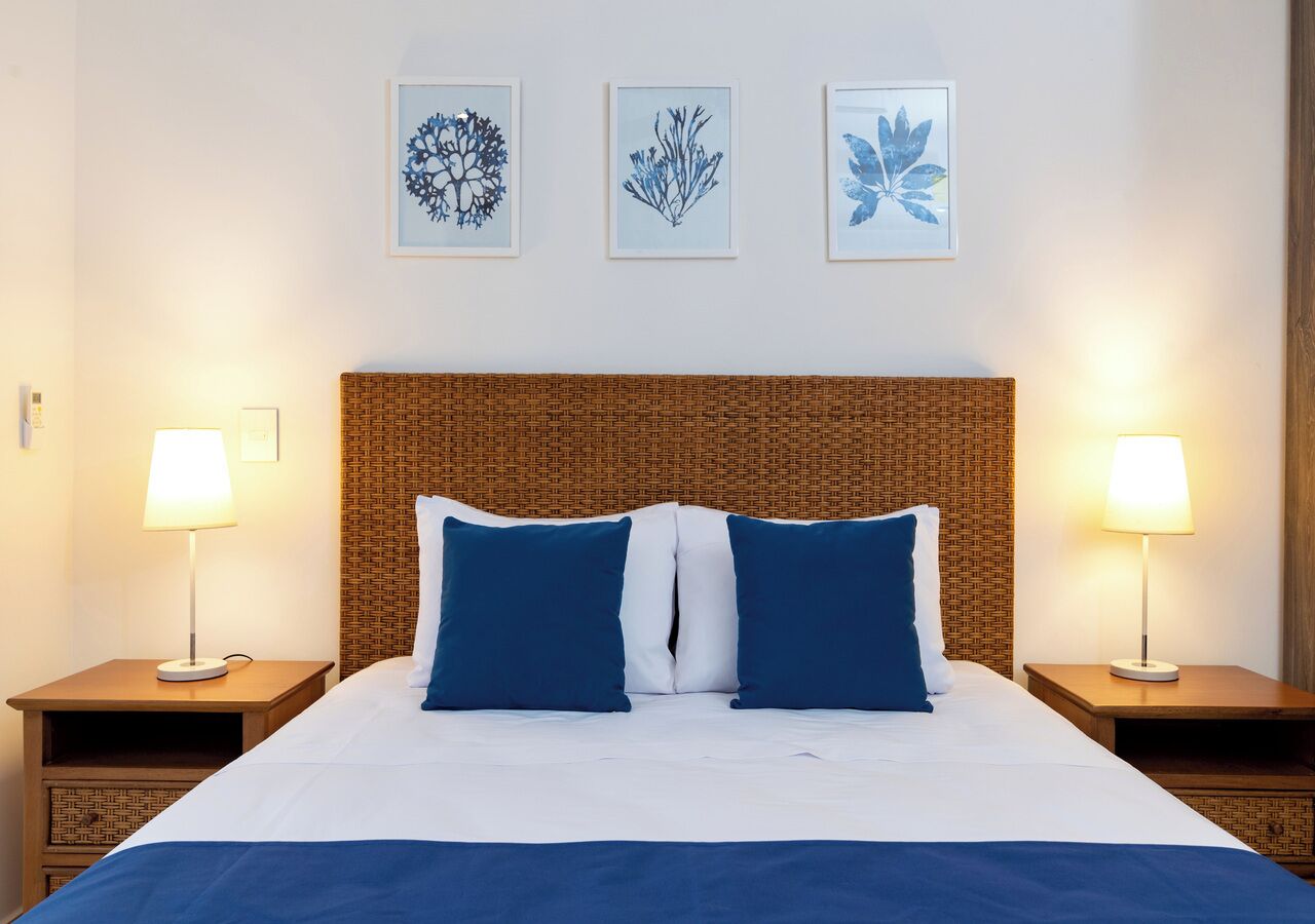 A hotel room with a bed, two bedside lamps, a woven headboard, and three framed wall art pieces.