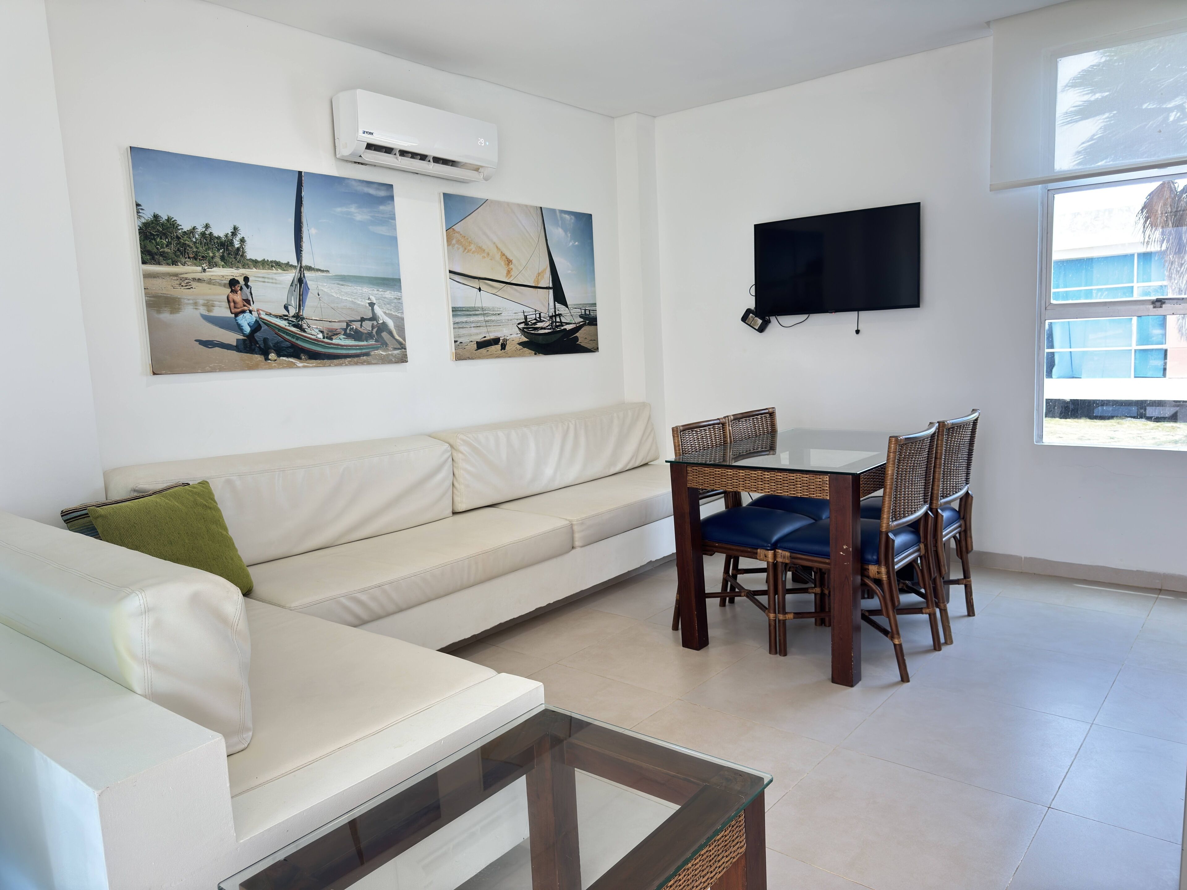 A modern living room with a white sofa, a glass-top coffee table, and a dining area with wicker chairs. There are two paintings on the wall depicting boats on the sea. An air conditioning unit is mounted above the sofa, and a flat-screen TV is on the wall.