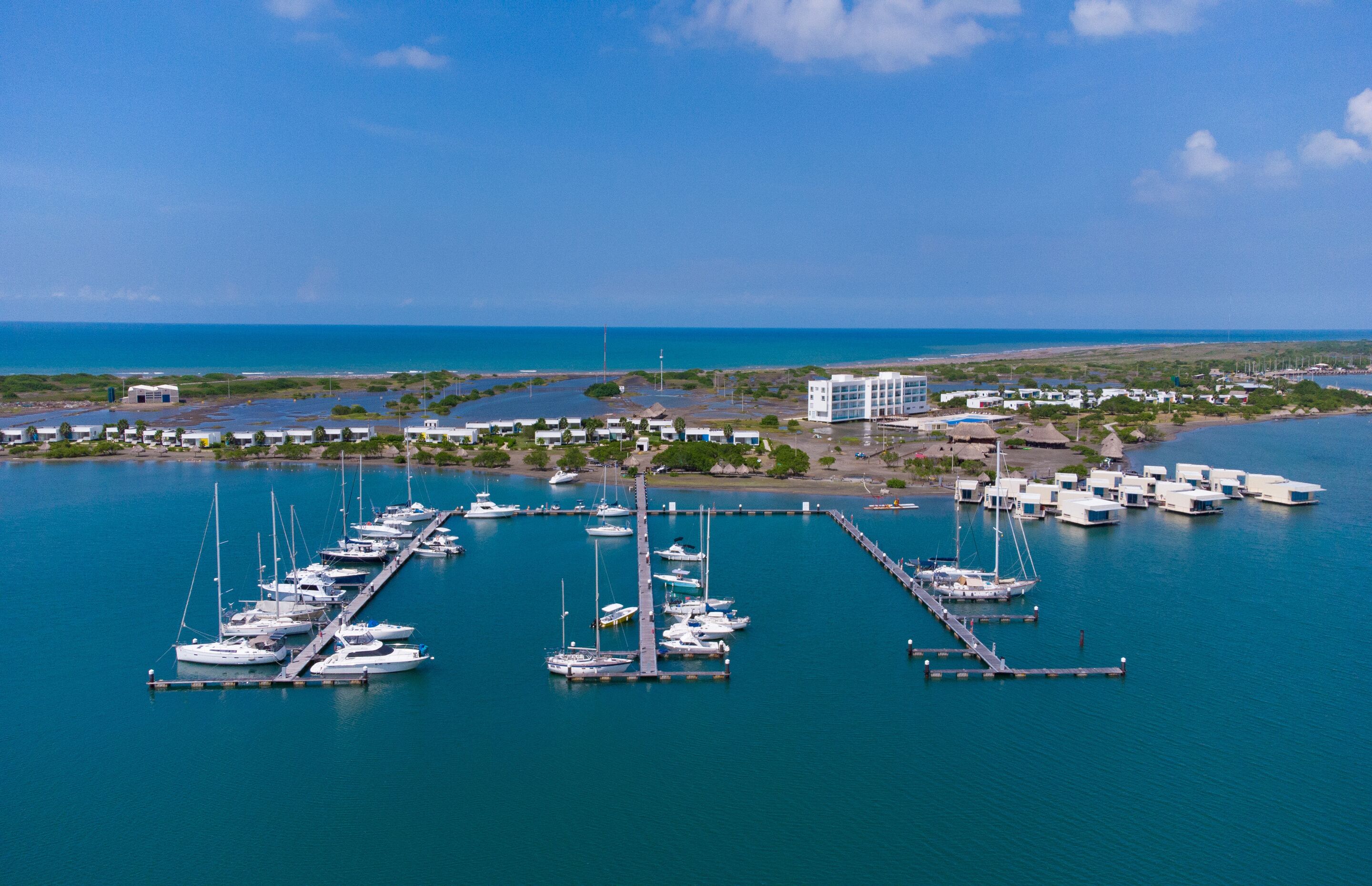 An aerial view of a marina with multiple boats docked, a large building, and a clear view of the ocean.