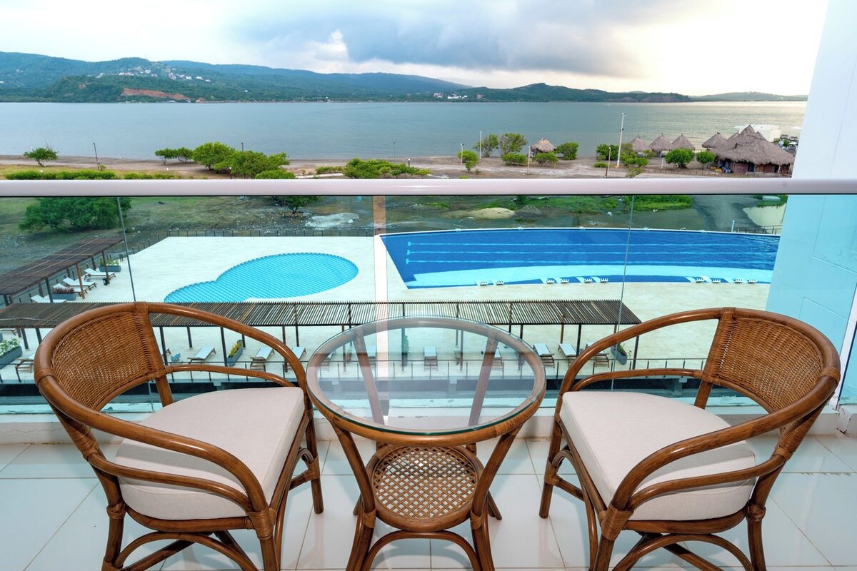 Un balcón con sillas de mimbre y una mesa de vidrio con vista a una piscina y un paisaje escénico de un lago y montañas.