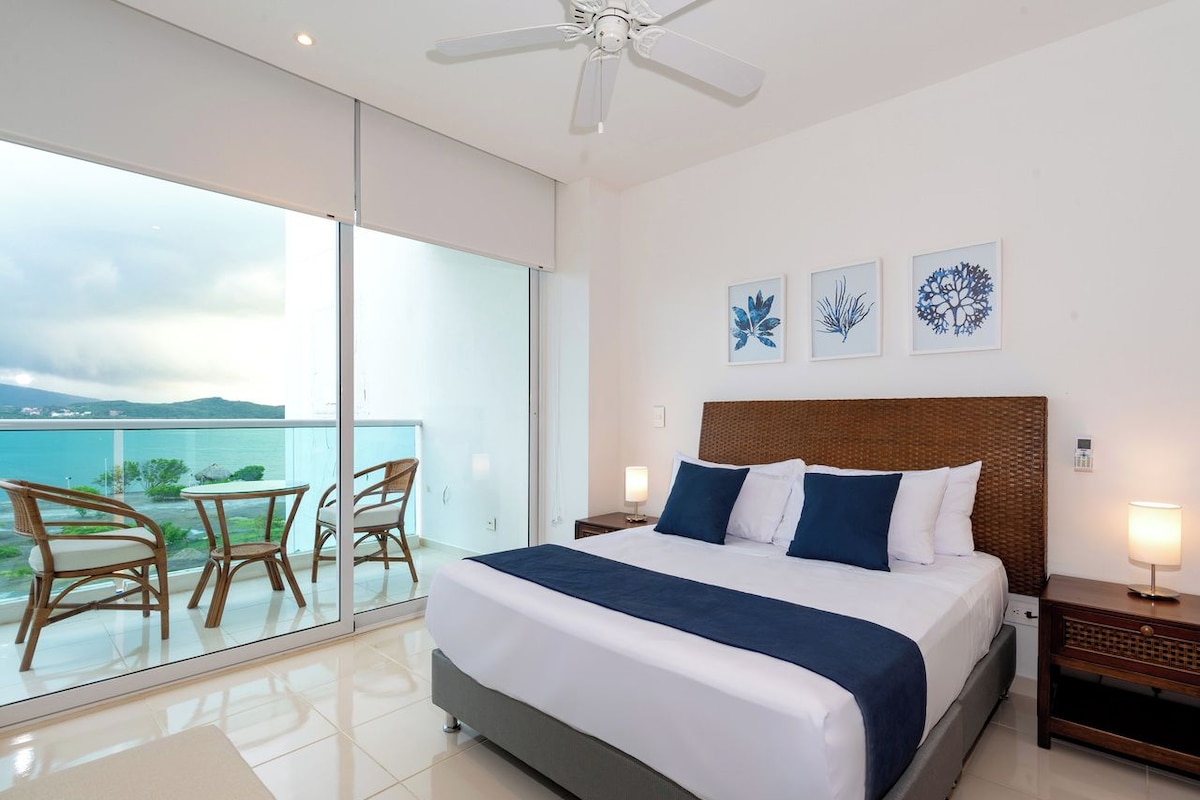 A bedroom with a large bed, a balcony with chairs, and artwork on the wall.