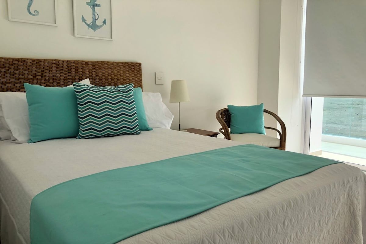 A neatly made bed with a teal bedspread and pillows, a wicker headboard, a bedside lamp, and a view of the sea through a window.