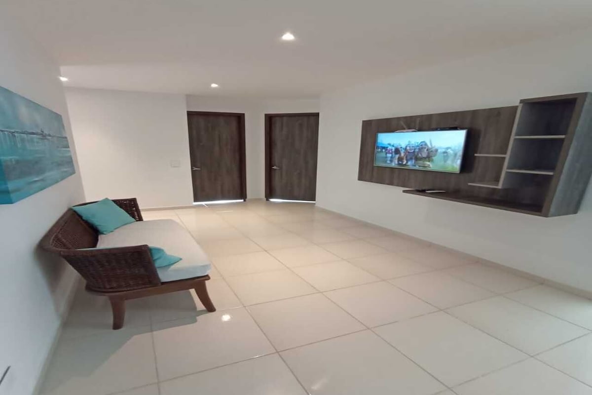 A spacious living area with a sofa, a flat-screen TV mounted on the wall, and a tiled floor.