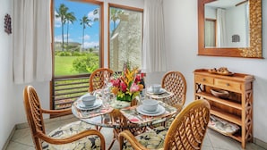A dining area with a glass table, wicker chairs, and a wooden cabinet with a mirror. - Kahala 712 Ground Floor By Suite Paradise (Koloa)