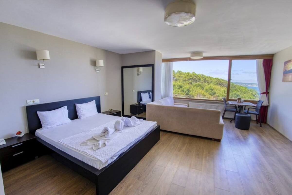 A modern hotel room with a large bed, a sofa, a dining table, and a view of a forested area.