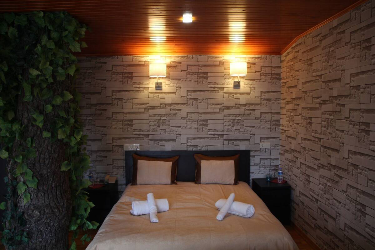 A hotel room with a brick-patterned wall, two bedside lamps, a bed with a beige bedspread, and a tree with green leaves on the left side.