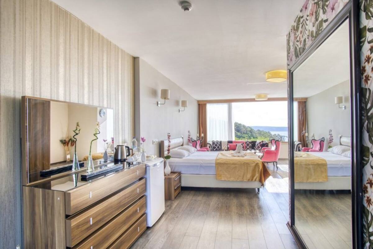 A modern bedroom with a large mirror, a wooden dresser, a bed with a view, and a small table with decorative items.