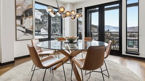 A modern dining area with a glass table, leather chairs, a unique chandelier, and large windows offering a mountain view. - The Lion Residences W602 (Vail)