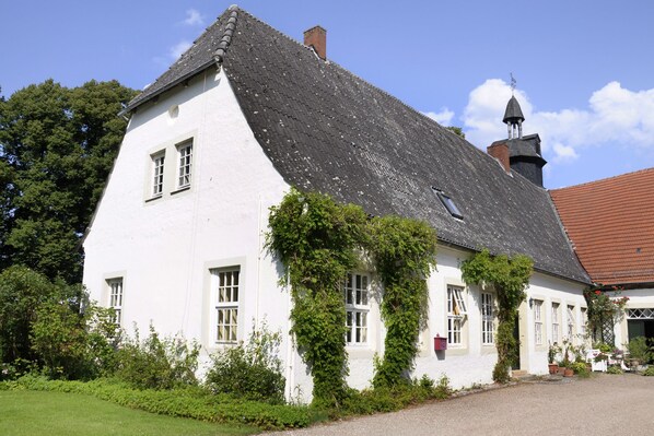 Een wit gebouw met een steil dak, muur begroeid met klimop en een kleine toren met windvaan.