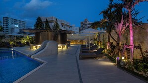 A pool area with a water slide, lounge chairs, and a stone wall with palm trees.
