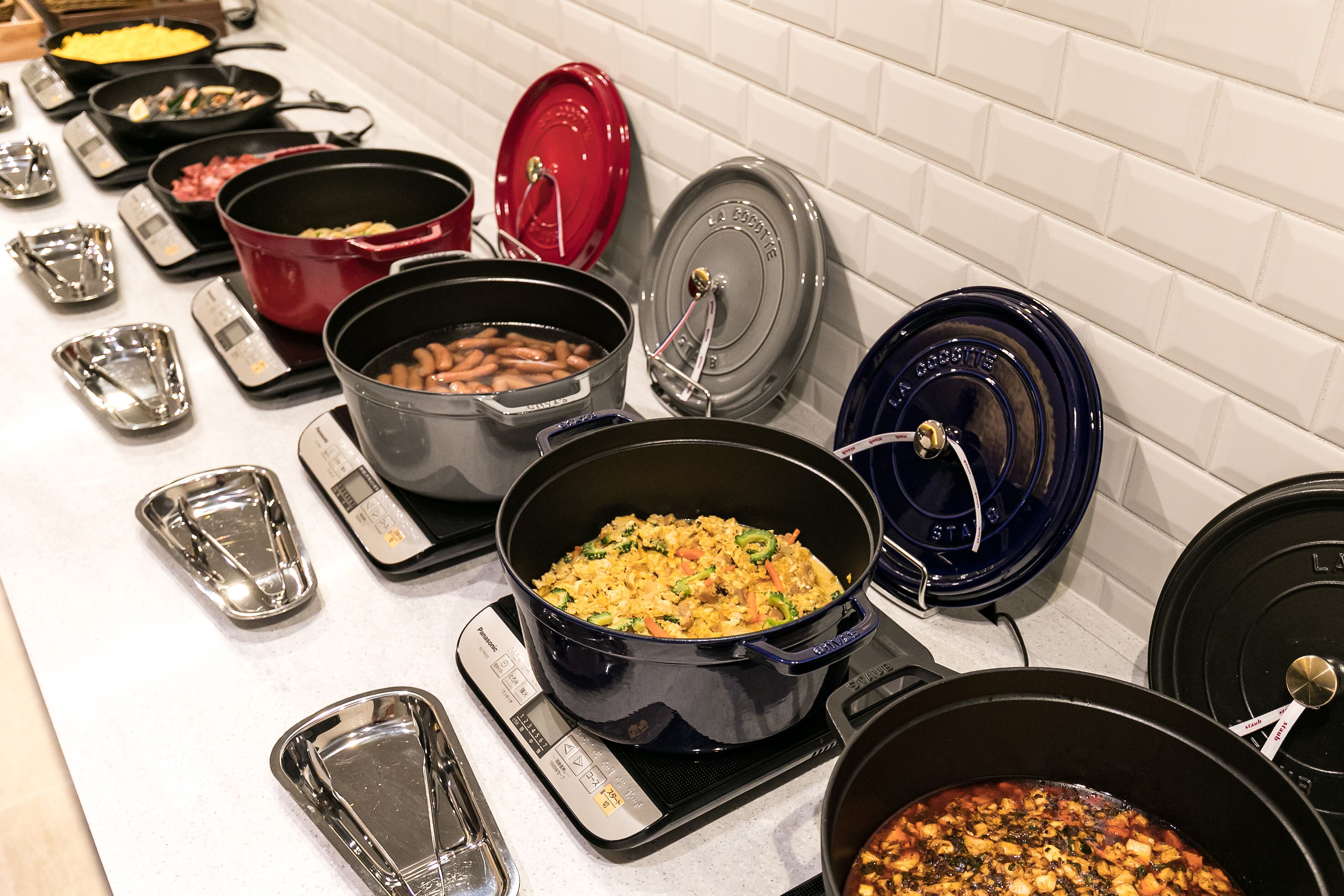 A buffet with various dishes in pots and pans, some with lids, on a counter with a white tiled backsplash.