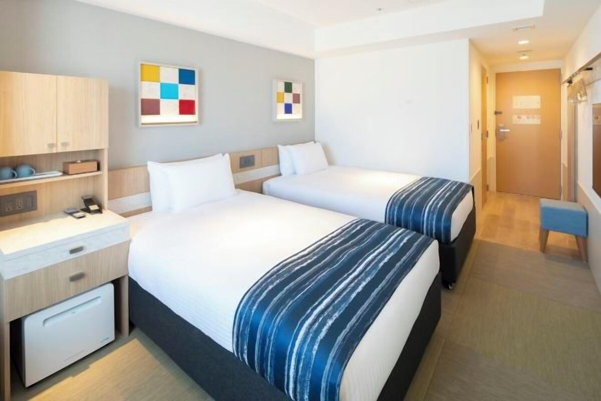 A hotel room with two beds, a desk, a chair, and a small table.