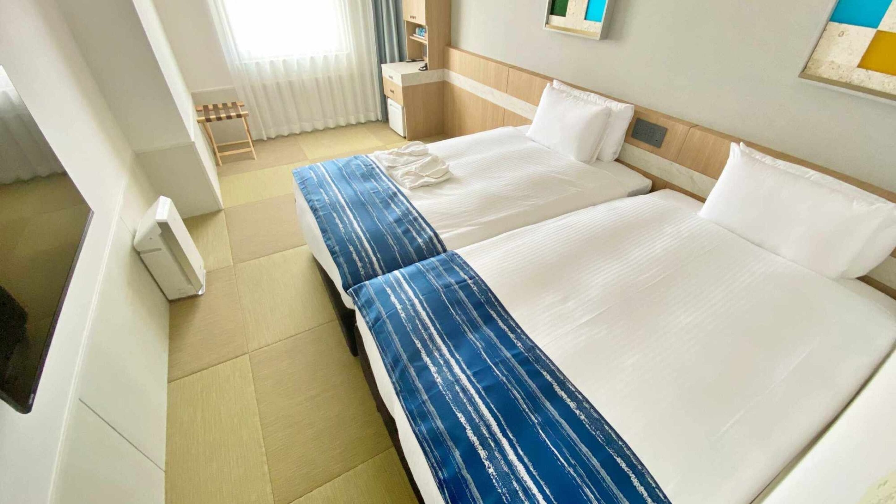 Ein Hotelzimmer mit zwei Betten, einem kleinen Schreibtisch, einem Stuhl und einem Fenster mit Vorhängen.
