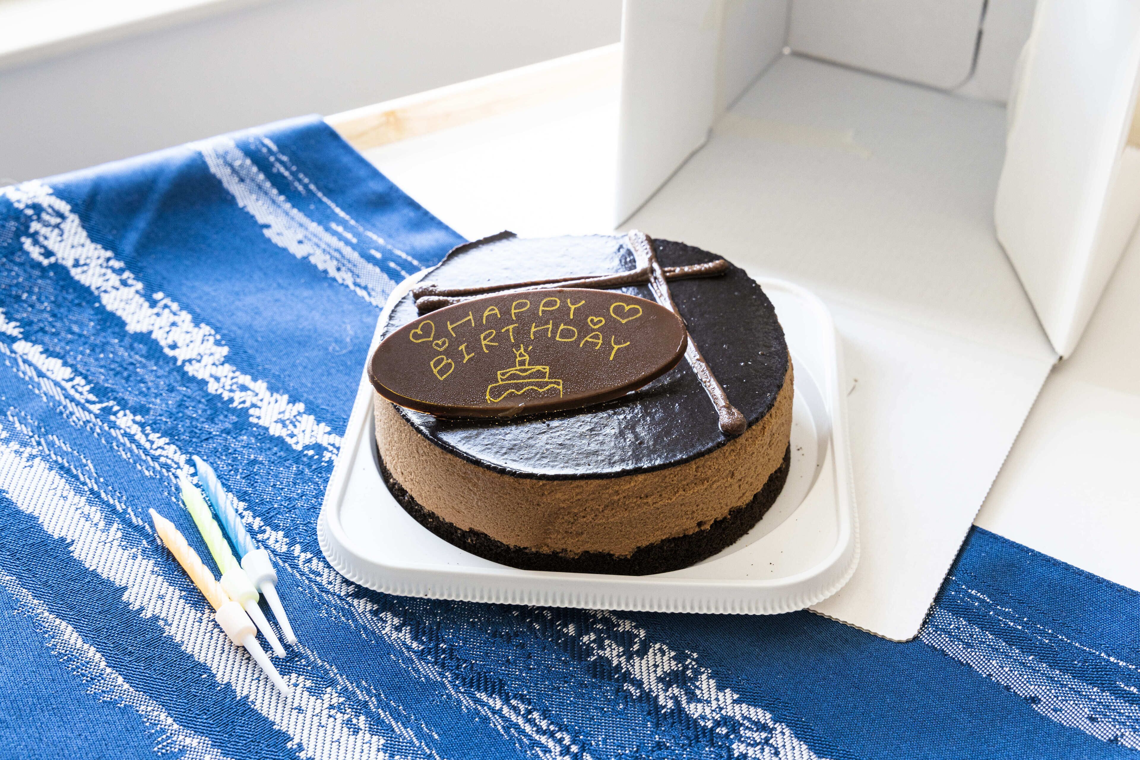 Un pastel de chocolate con glaseado que dice 'Happy Birthday' sobre un pedestal blanco sobre un mantel azul y blanco a rayas.