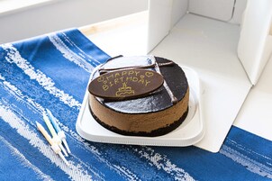 Un pastel de chocolate con glaseado que dice 'Happy Birthday' sobre un pedestal blanco sobre un mantel azul y blanco a rayas.