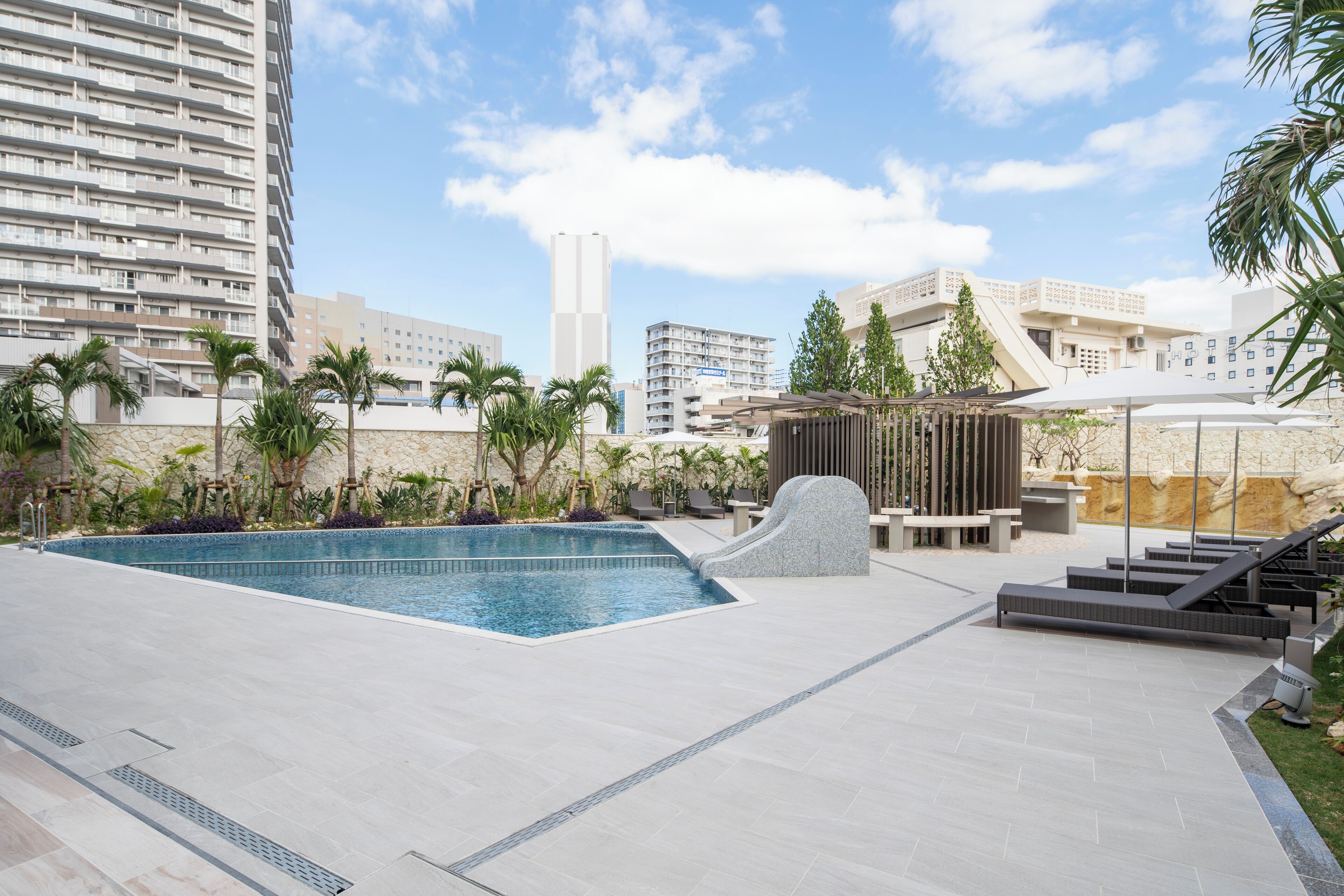 A modern outdoor pool area with a stone waterfall feature, surrounded by palm trees and a cityscape in the background.