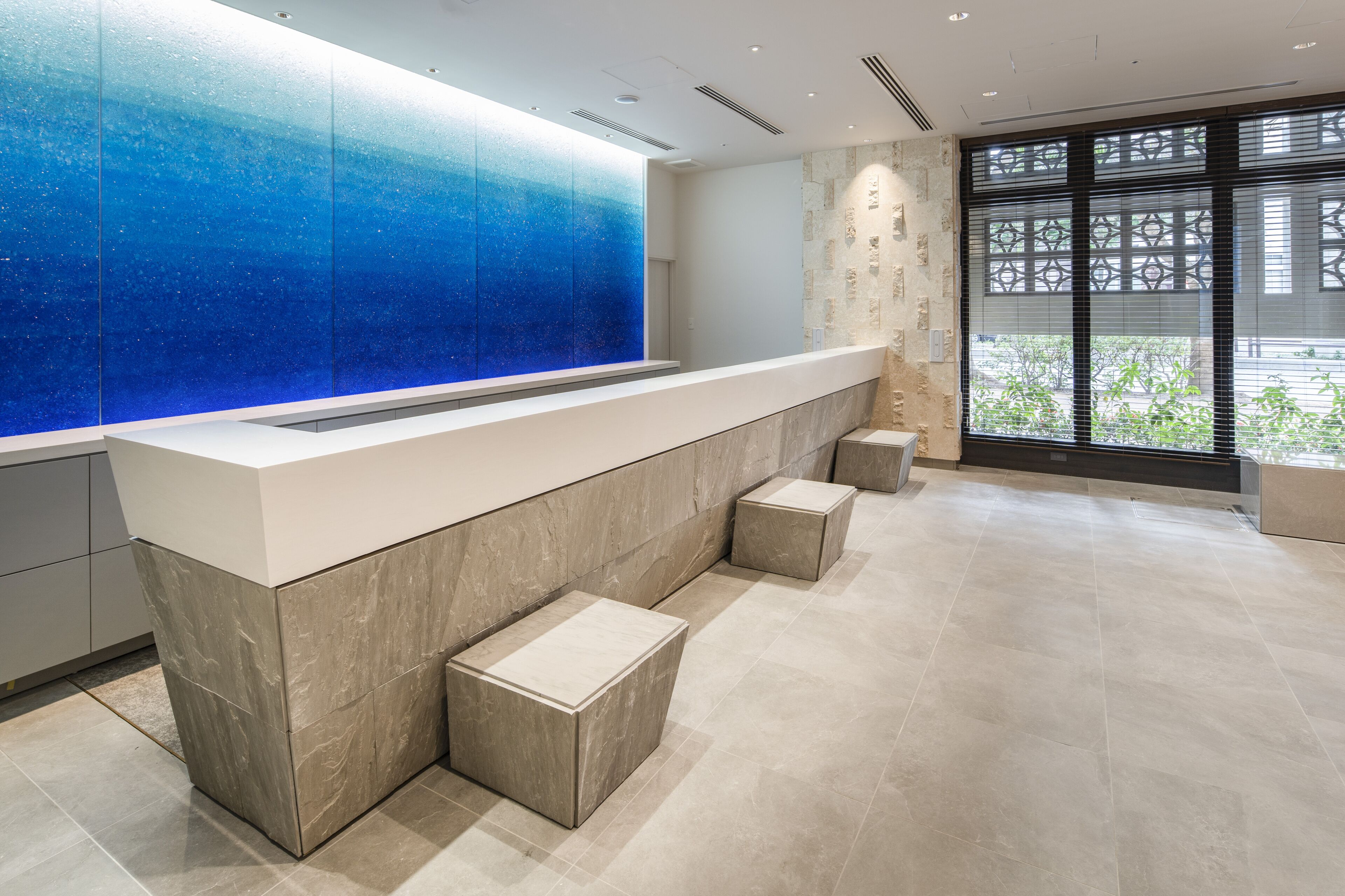 A reception area with a modern design, featuring a marble counter, a blue glass wall, and a view of greenery outside.