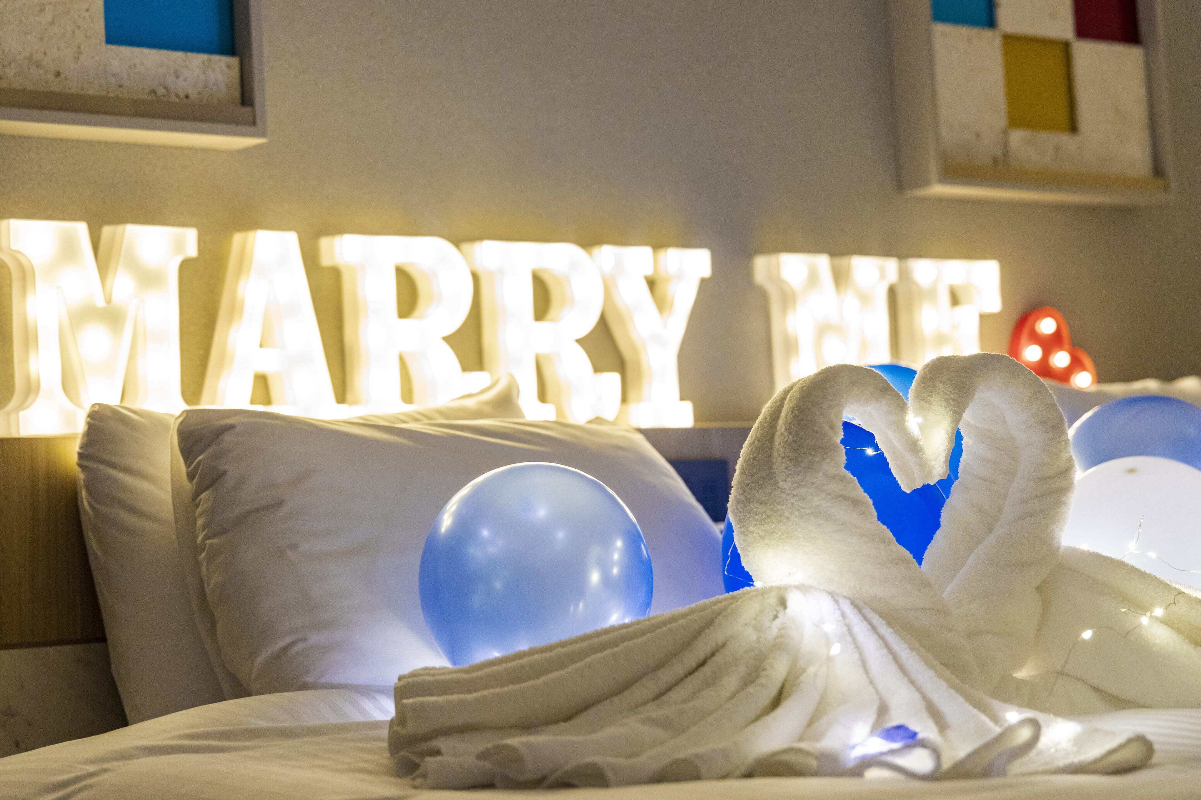 A bed with white linens, a heart-shaped towel, and illuminated 'MARRY ME' letters in the background.