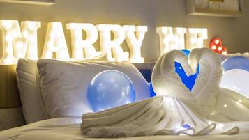 A bed with white linens, a heart-shaped towel, and illuminated 'MARRY ME' letters in the background.