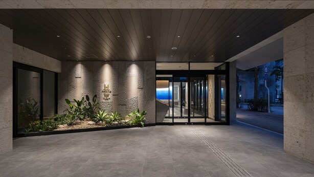 A modern building entrance with a stone facade, glass doors, and a landscaped area with plants.