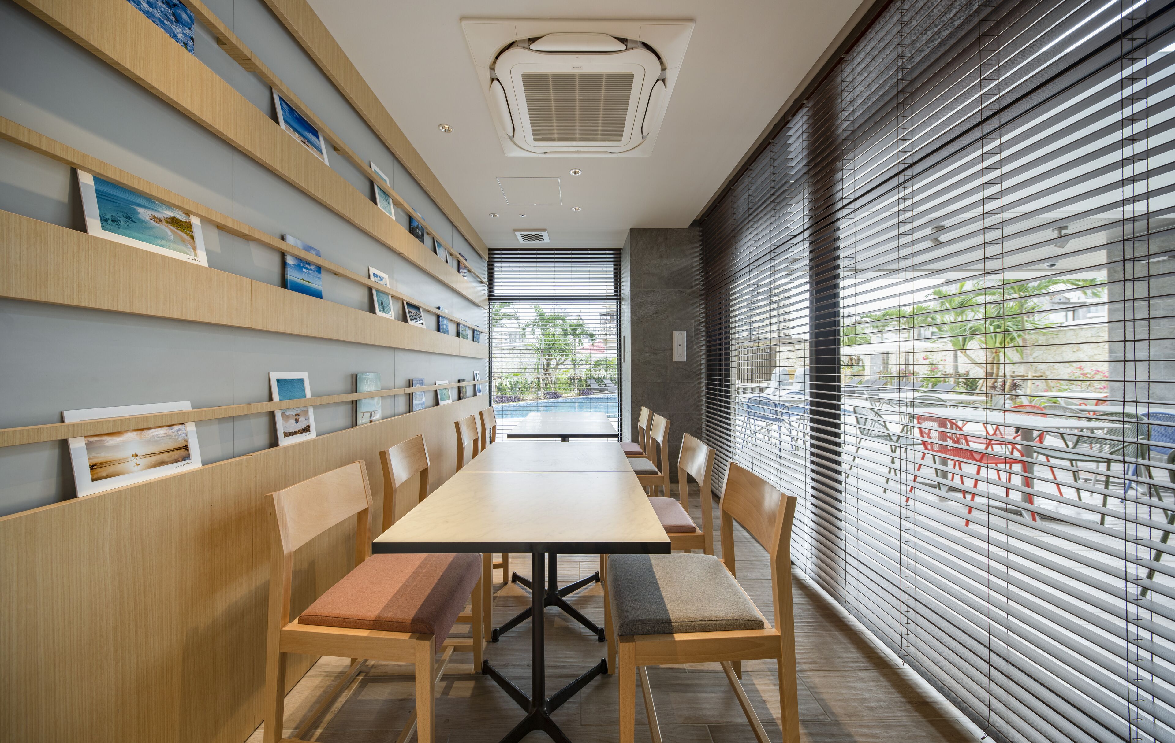 A modern restaurant with a long wooden table and chairs, a wall-mounted shelf with framed pictures, and a large window with blinds.