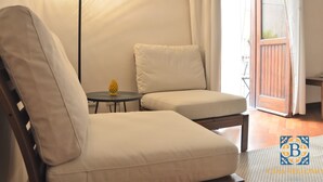 A room with a wooden-framed bed, a white armchair, a small round table with a yellow object, and a view of the outside through a glass door.