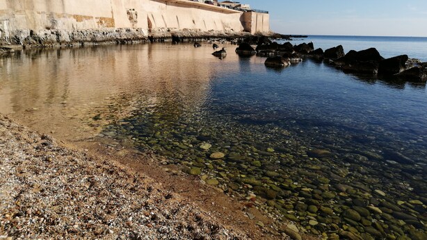A coastal scene with a rocky shoreline, clear water, and buildings along the edge.