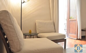 Une pièce avec un lit à cadre en bois, un fauteuil blanc, une petite table ronde sur laquelle se trouve un objet jaune, et une vue sur l’extérieur par une porte vitrée.