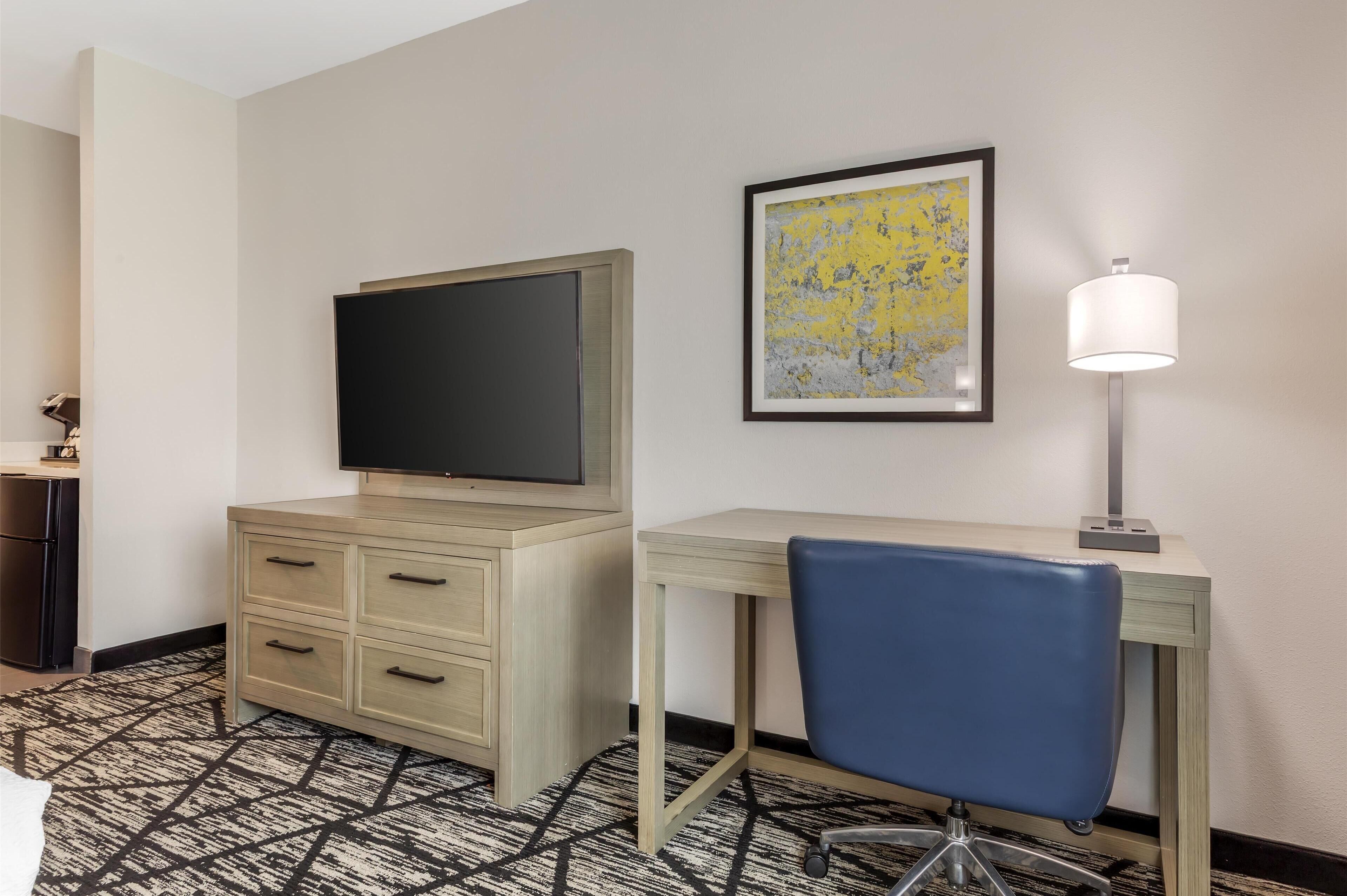 A hotel room with a bed, a desk with a chair, a television, a framed artwork, and a floor lamp.