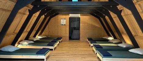 Une chambre située sous les combles, avec des poutres apparentes, quatre lits simples, un ventilateur de plafond et un radiateur.