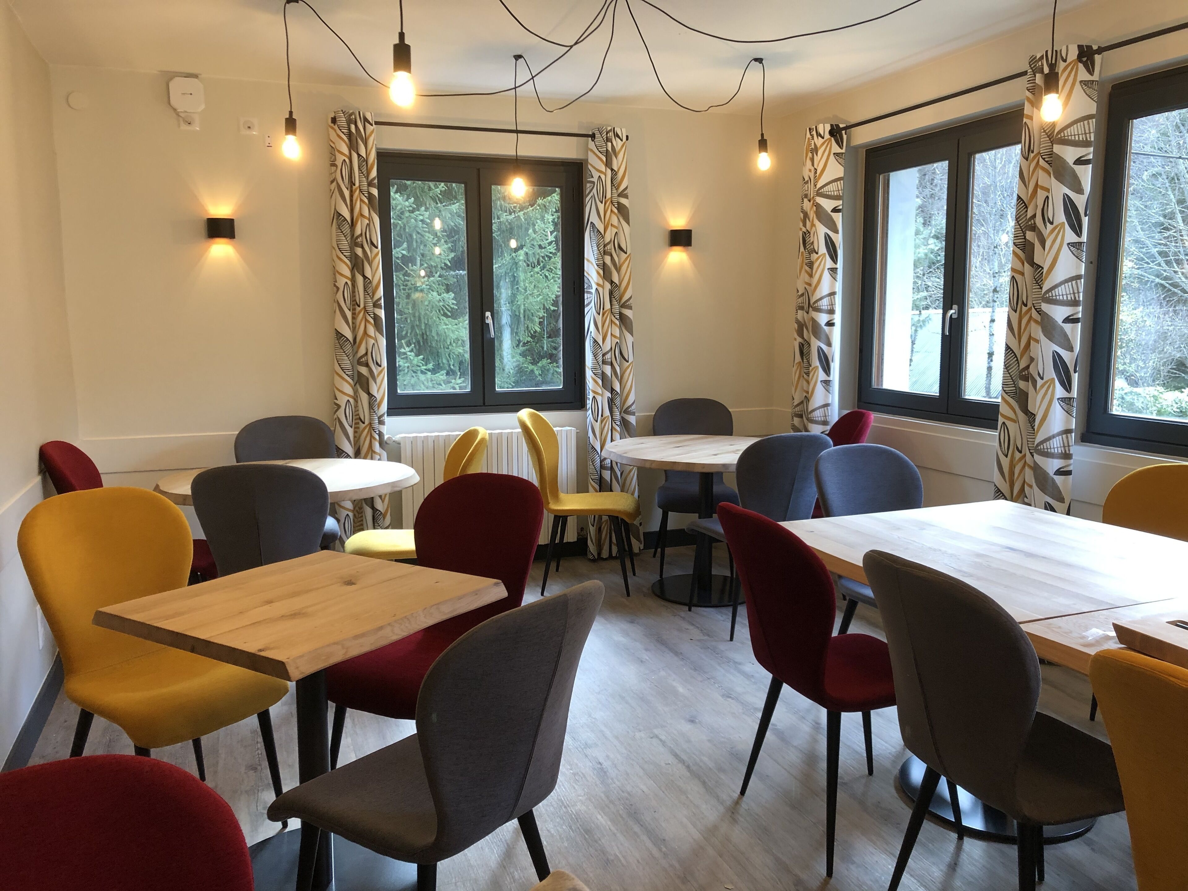 Un comedor con mesas y sillas redondas, ventanas grandes con cortinas y bombillas colgantes.