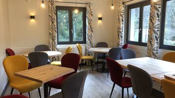 Un comedor con mesas y sillas redondas, ventanas grandes con cortinas y bombillas colgantes.