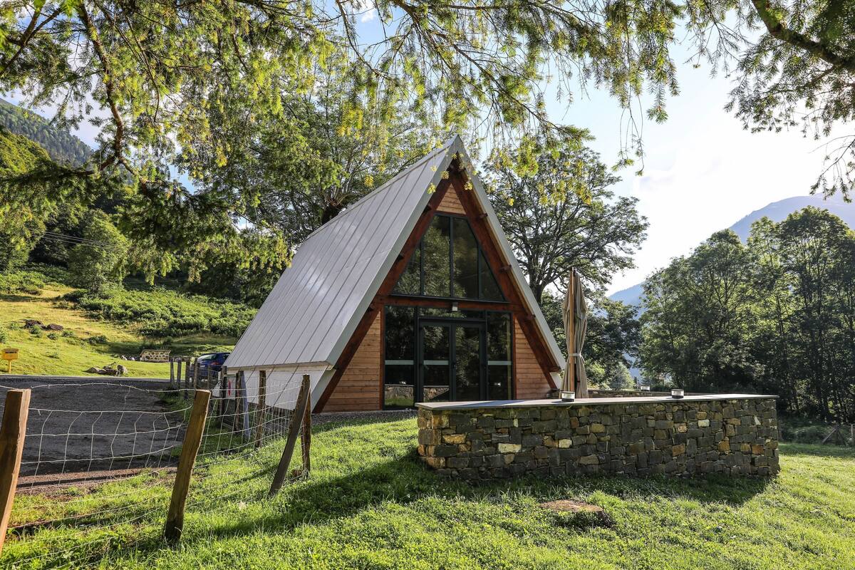A small, triangular-roofed cabin with a stone foundation, surrounded by lush greenery and trees.