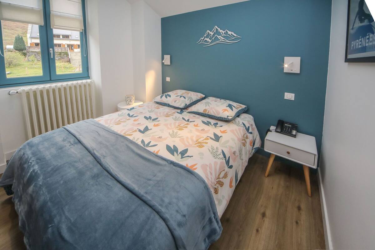 A bedroom with a bed, a bedside table, a wall-mounted light, and a window with a view of the outside.