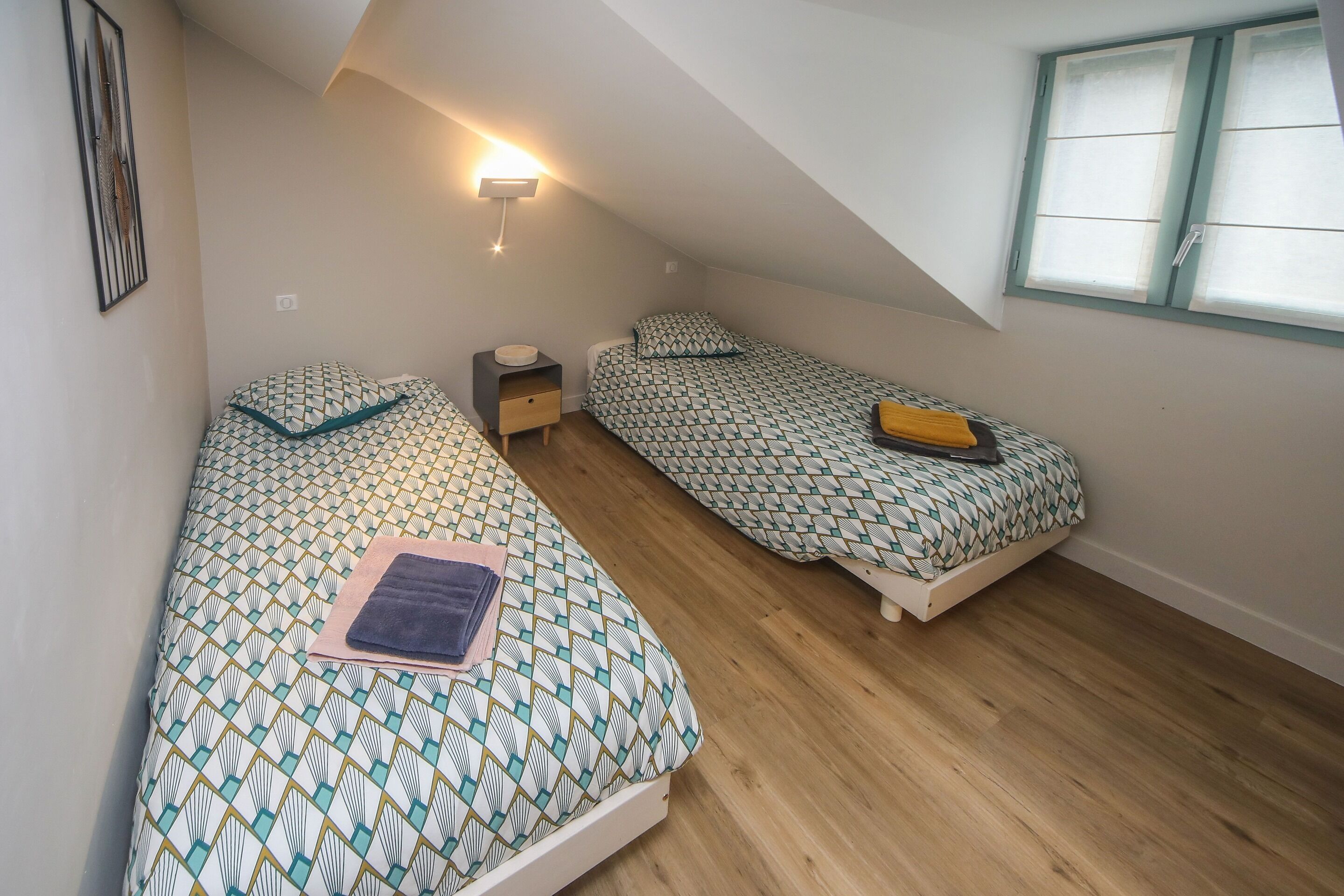 A small, cozy room with two single beds, a nightstand, a wall-mounted light, and a window with curtains.