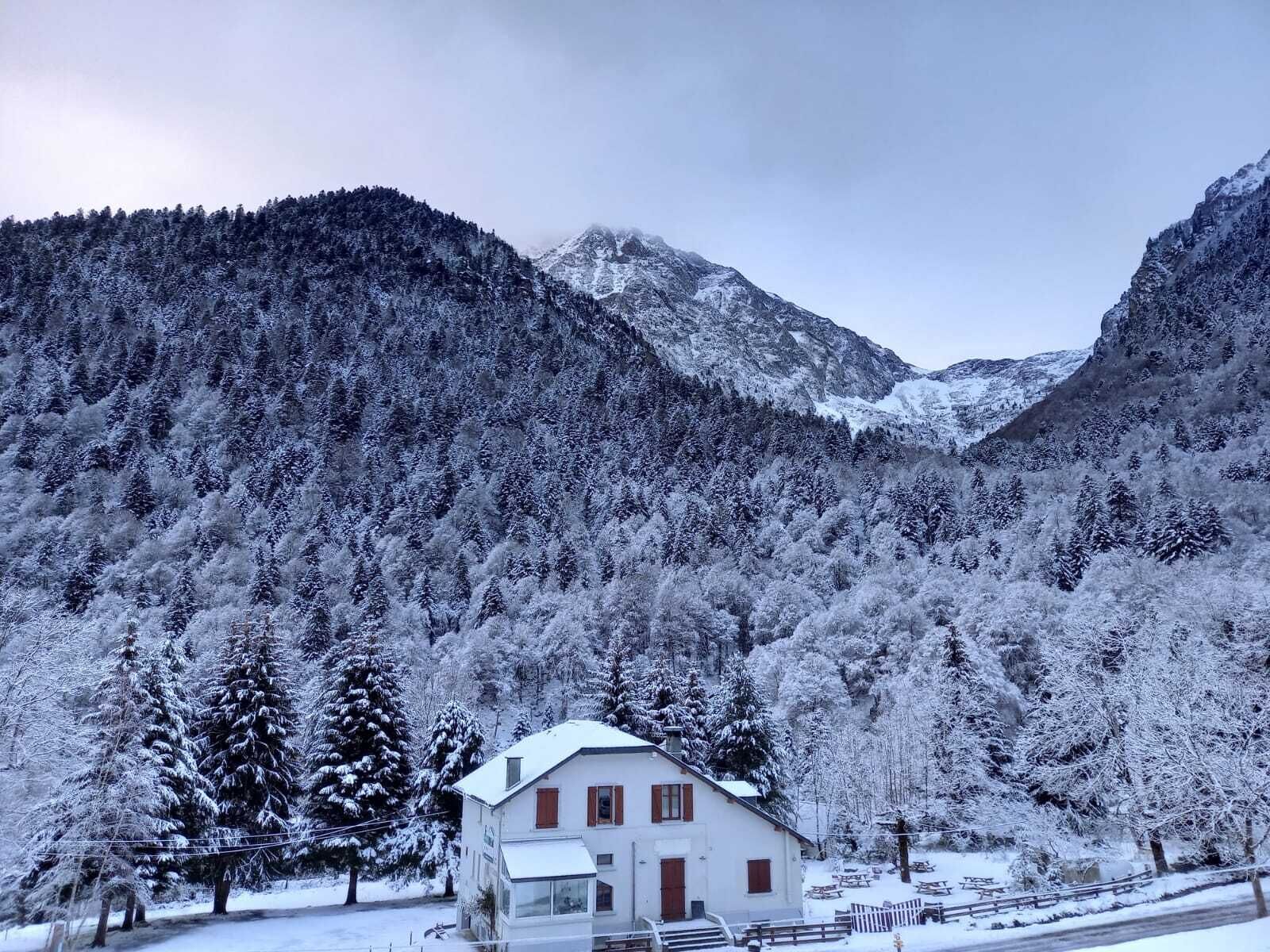 Un edificio blanco con techo inclinado, rodeado de árboles y montañas cubiertas de nieve.