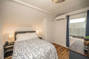 Quarto com cama, abajures, relógio, janela com cortinas e uma pequena planta sobre uma mesa de madeira.
