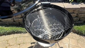 Una barbacoa amb tapa i reixa metàl·lica, col·locada en un pati pavimentat amb un mur de pedra i vegetació al fons.
