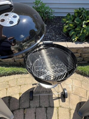 A barbecue grill with a lid and a wire rack, placed on a paved patio with a stone wall and greenery in the background.