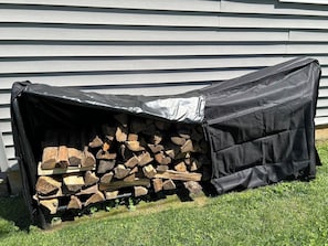 A stack of firewood covered with a black tarp, placed on grass in front of a building with horizontal siding.