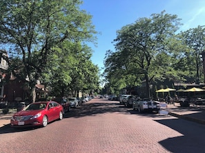 A paved street lined with parked cars and trees, with a brick sidewalk and outdoor seating area with umbrellas.