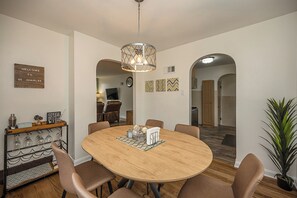 Un coin repas comprenant une table ronde en bois, six chaises, un luminaire suspendu et un espace dédié au café, avec des verres et un panneau indicateur.