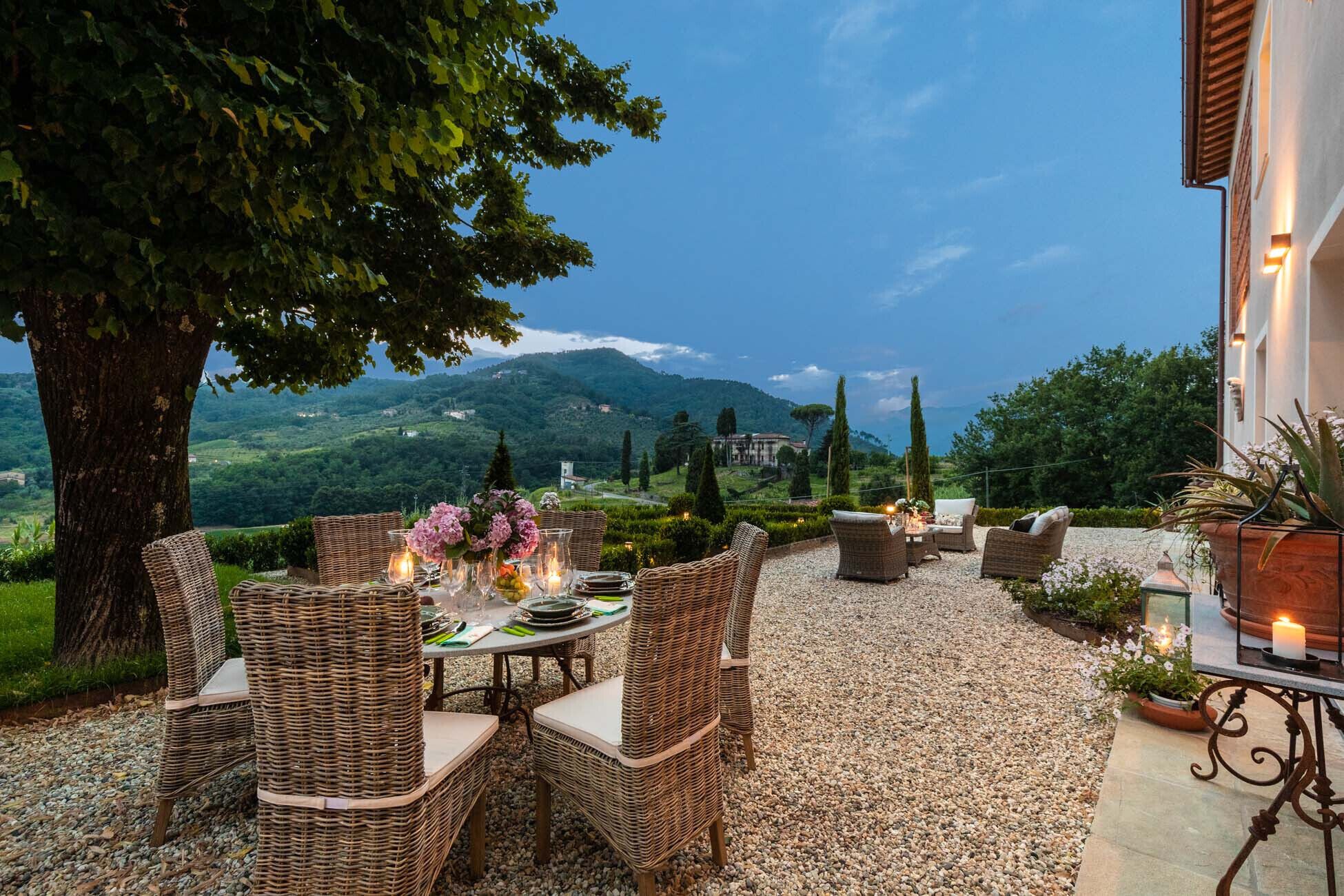 An outdoor dining area with wicker furniture, a table set with plates, glasses, and candles, surrounded by a gravel path and greenery.