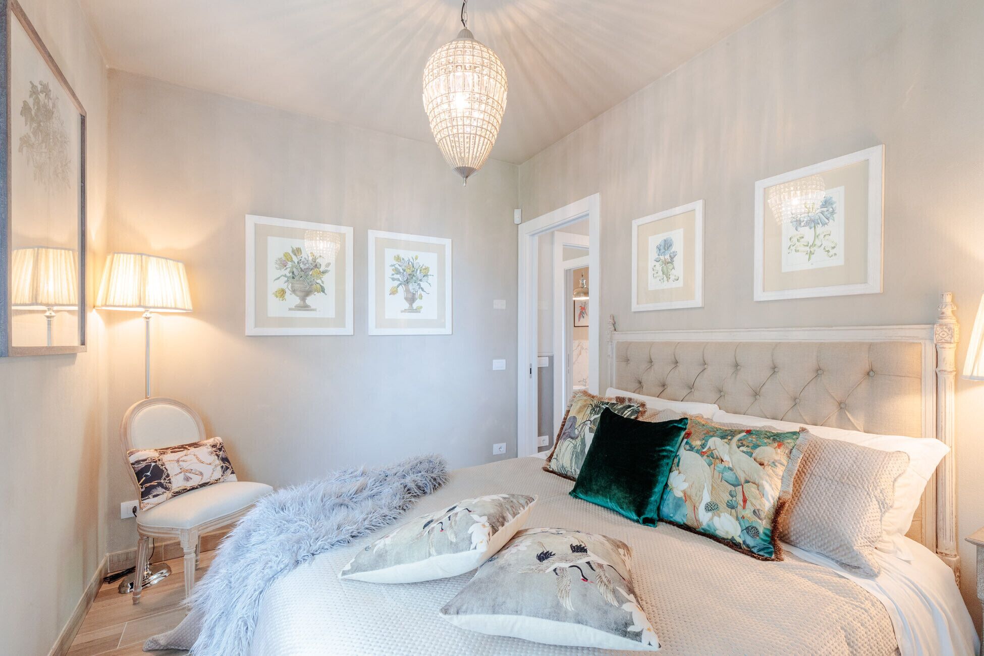 A bedroom with a large bed, a chandelier, framed floral artwork, and a chair.