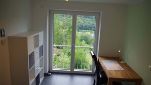 Ein Zimmer mit einem Holztisch, einem Stuhl, einem Schreibtisch, einem Bücherregal und einer Glastür, die auf einen Balkon mit Blick ins Grüne führt.