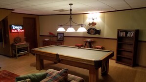 Una mesa de billar en una sala con un letrero de neón que dice "Doug's Pool Room", una estantería de madera y un candelabro.