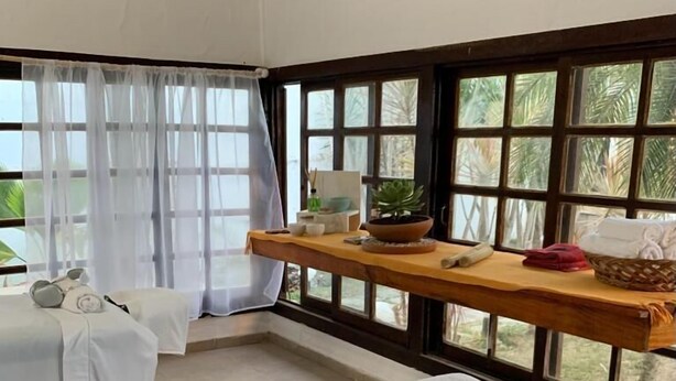A room with two beds, a wooden shelf with toiletries, and large windows with a view of greenery. - Costa del Sol Pousada (Búzios)