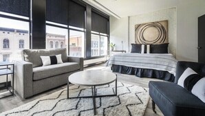 A modern hotel room with a large bed, two sofas, a round coffee table, and a view of the city through large windows. - Cassius Quarters@The713! Downtown, large, upscale suite w/private balcony (Louisville)